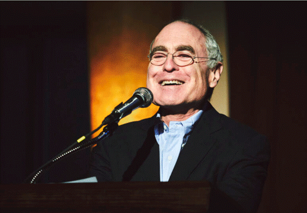 A person in a suit and glasses speaking into a microphone
Description automatically generated with low confidence
