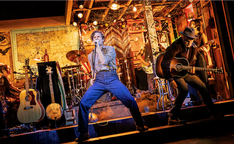 A person singing into a microphone on a stage with a person playing guitar
Description automatically generated