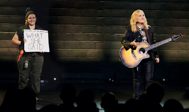 A person holding a guitar and a person holding a sign
Description automatically generated