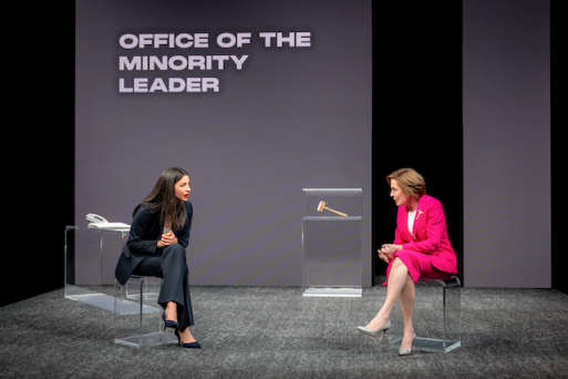 Two women sitting on chairs in front of a screen Description automatically generated