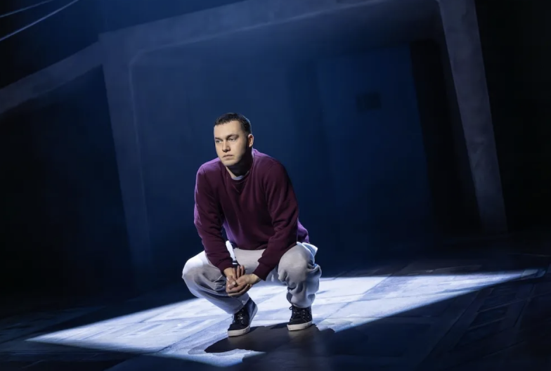 A person in a purple sweater squatting on a stage Description automatically generated