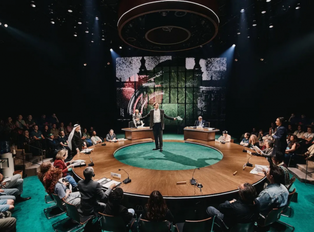 A person standing on a round table with people sitting in chairs

Description automatically generated